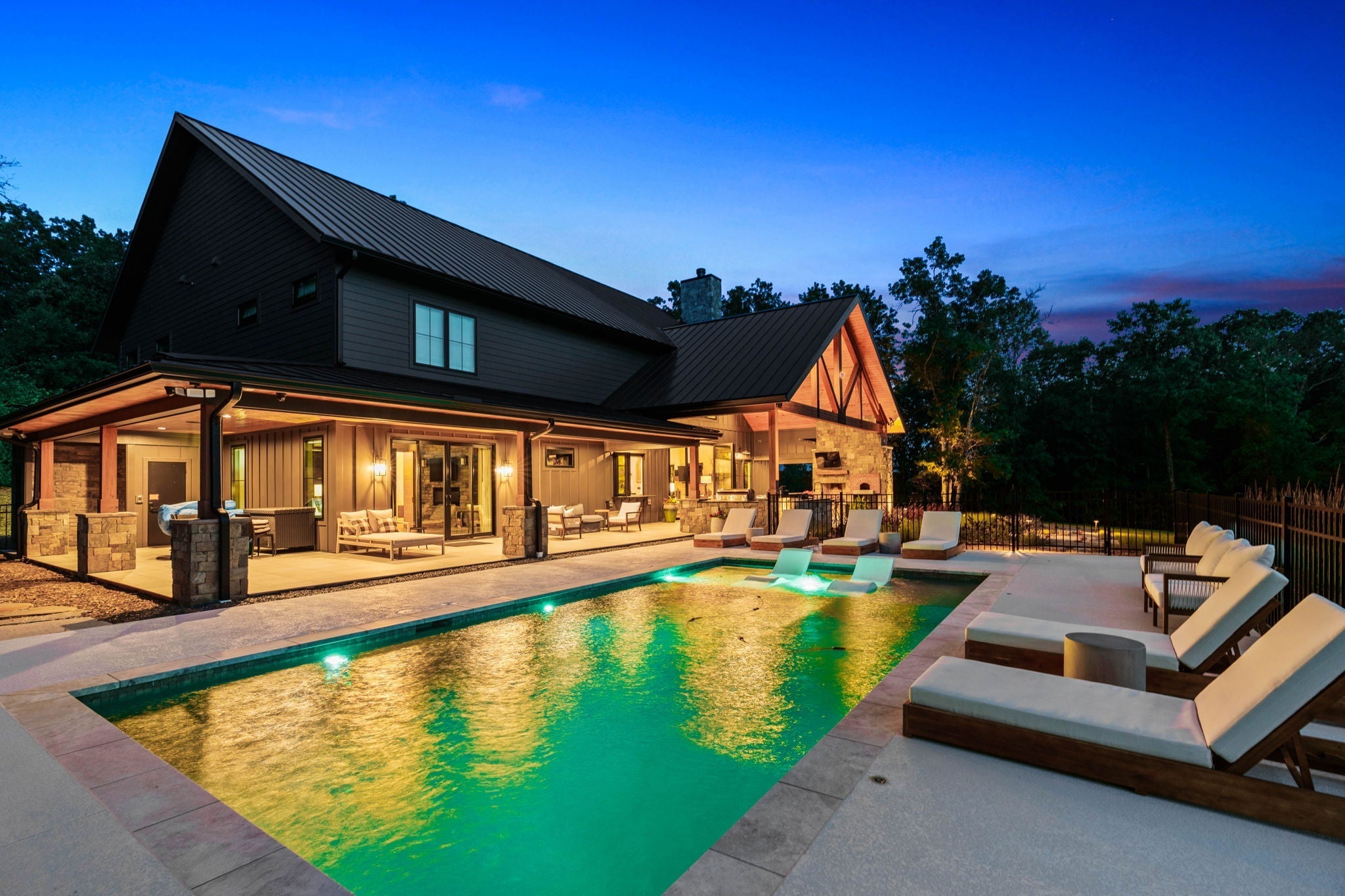 Modern barndominium with black metal roof, board-and-batten siding, covered patio, and in-ground pool lit at dusk
